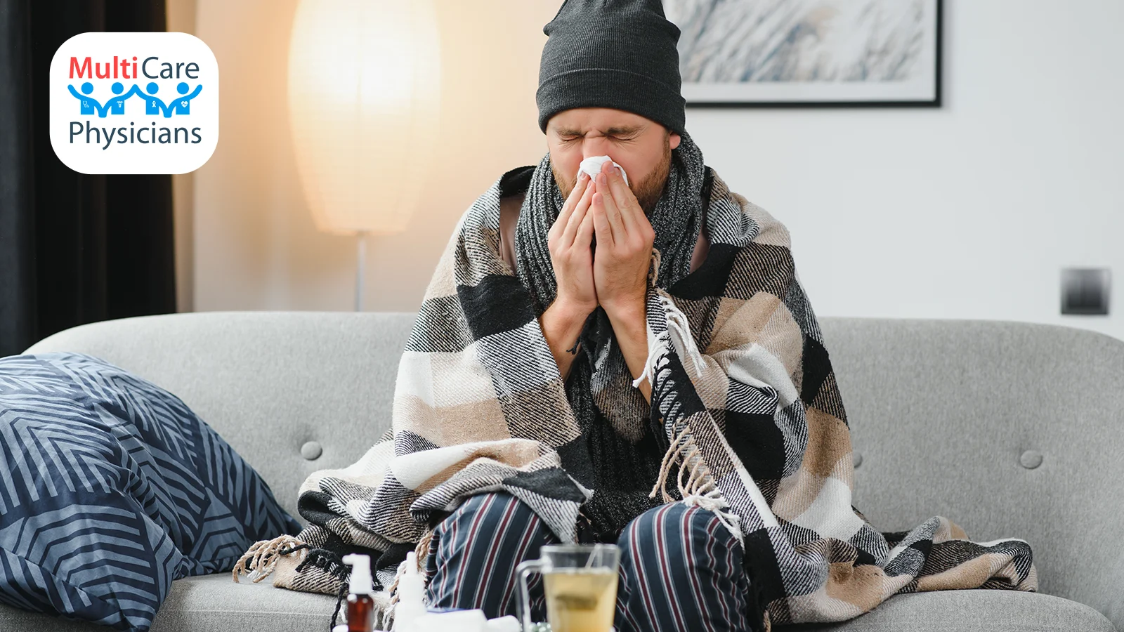 A sick man wrapped in a warm blanket sits on a couch, blowing his nose, appearing to suffer from flu symptoms. He is surrounded by flu remedies, including tea and medicine. The MultiCare Physicians logo is visible in the top left corner, reinforcing a healthcare and wellness message about flu prevention and treatment.