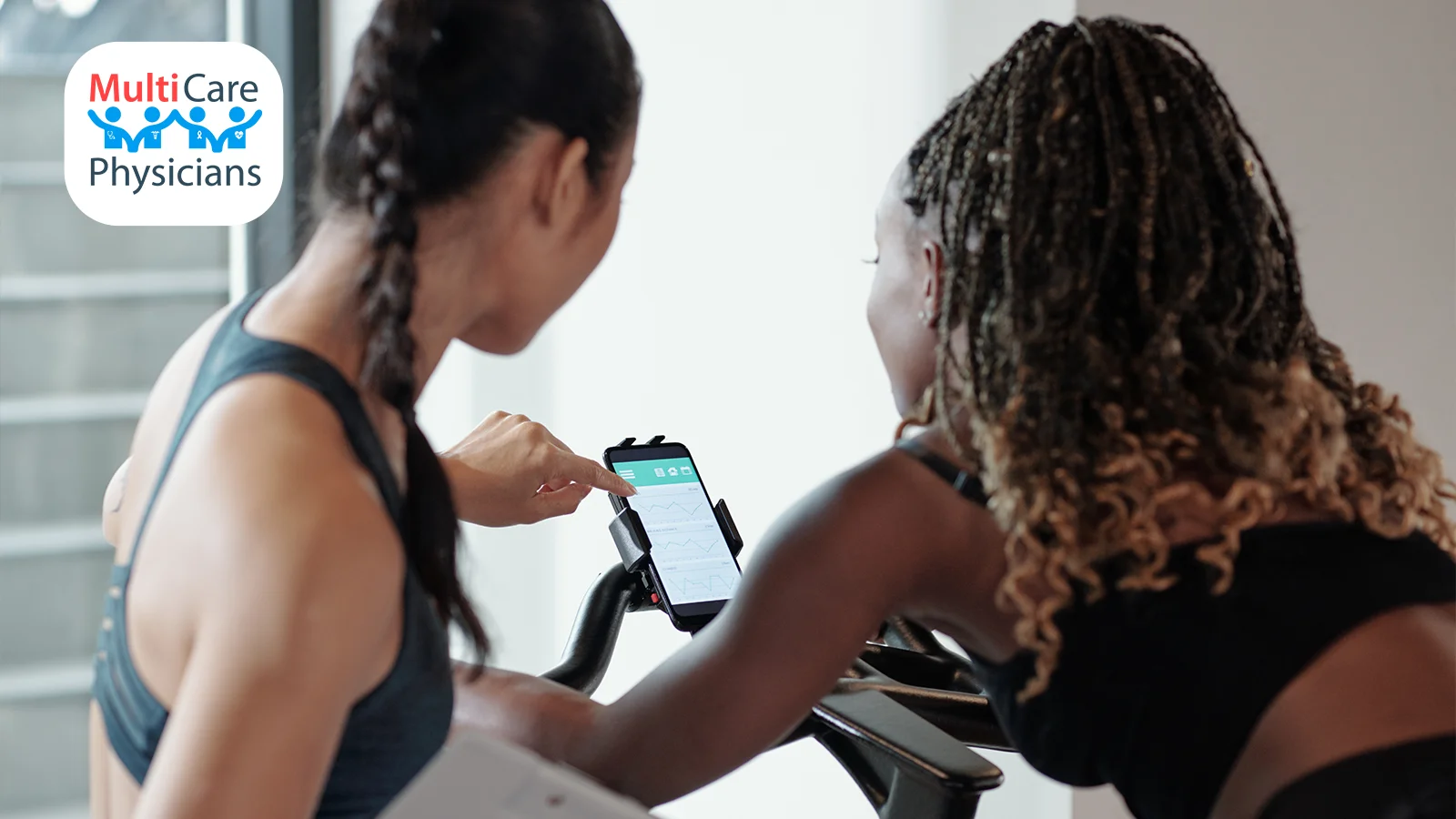 Two women in athletic wear using a stationary bike while reviewing fitness data on a smartphone. One woman points at the screen, highlighting performance metrics from a workout app. The image represents the role of workout apps in tracking fitness progress and improving exercise routines.